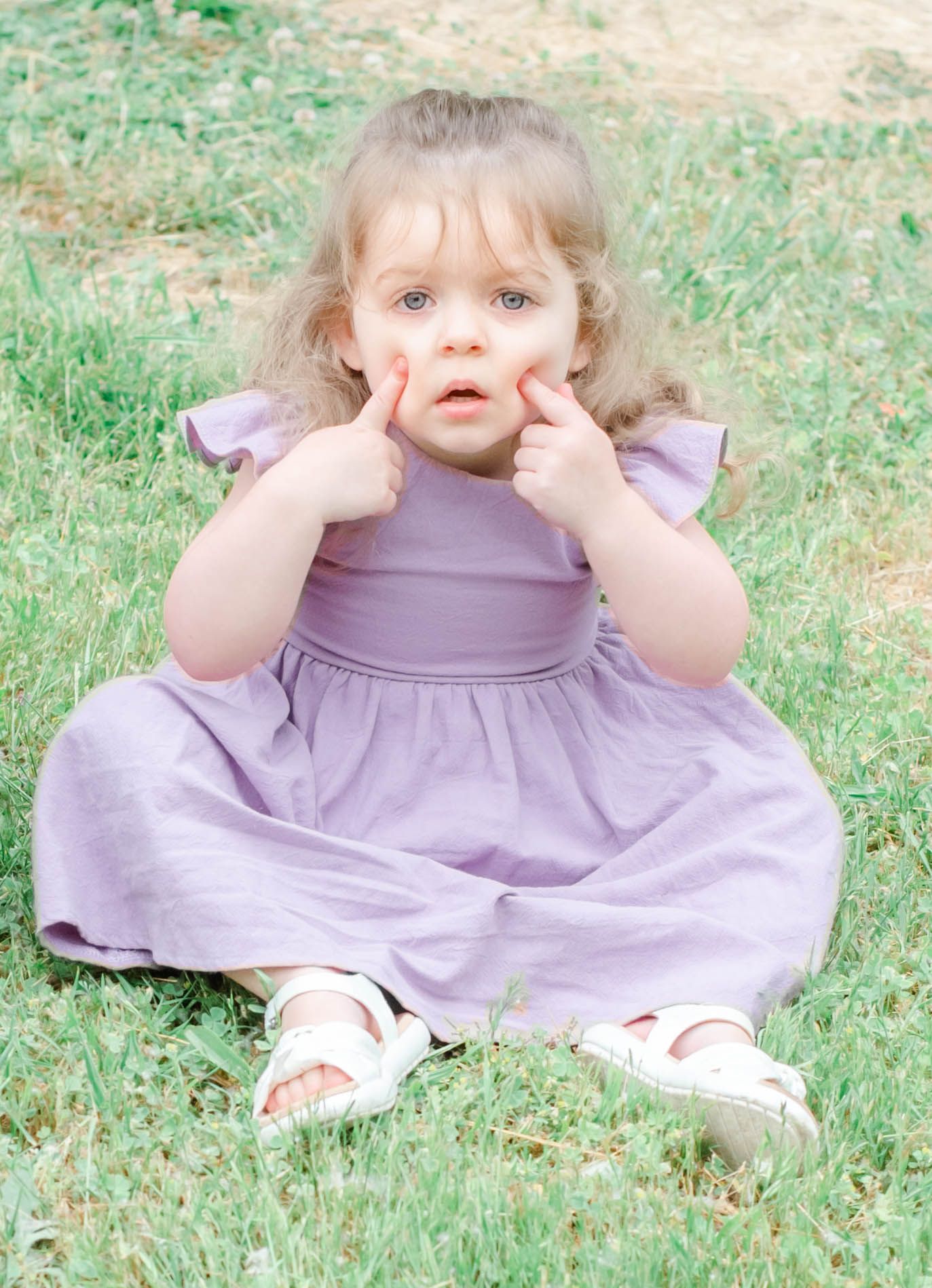 Young child in lavender dress sitting on grass playing peek-a-boo.