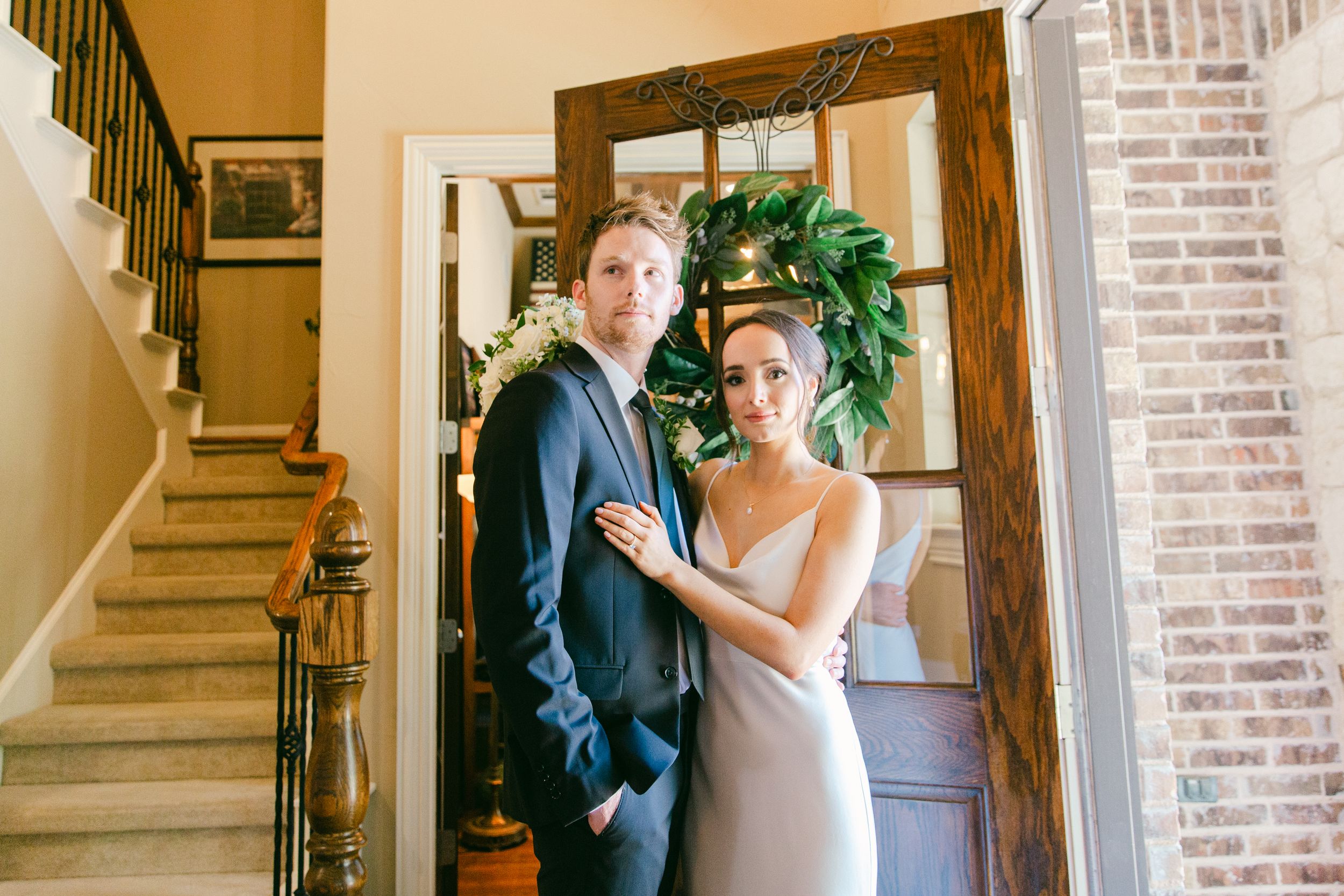 Couple in formal wedding attire poses by wooden staircase and brick wall.