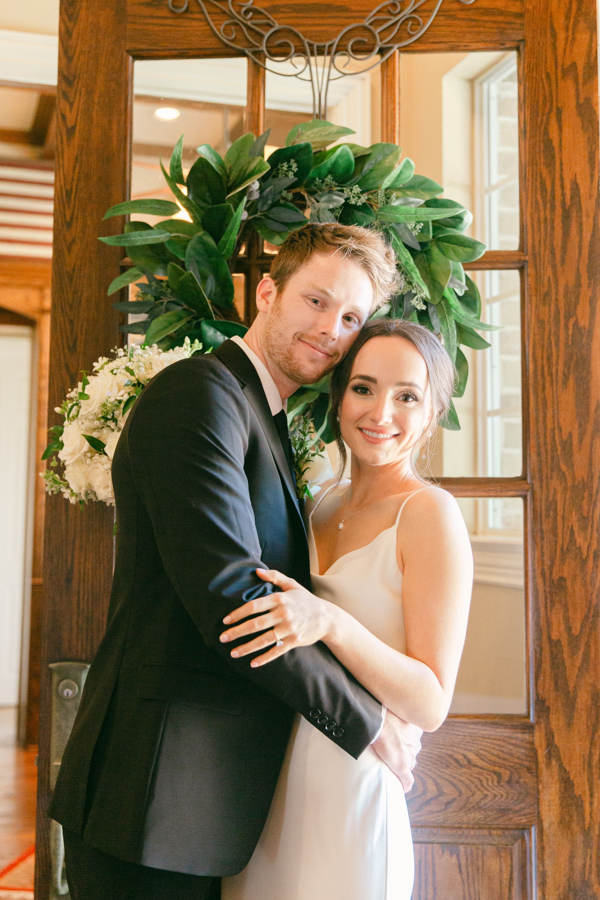 Newlyweds pose under elegant green wreath in wooden doorframe.