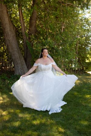 Bride in white off-shoulder ball gown twirling on green grass beneath large trees  at Headlands Park, Mackinaw City, MI