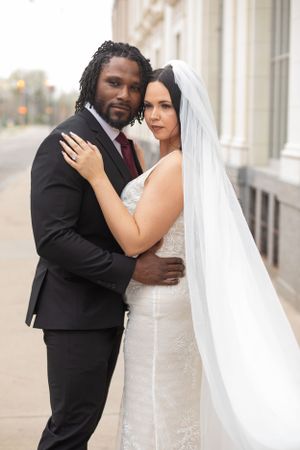 A couple embraces in formal wedding attire against a classic architectural backdrop.