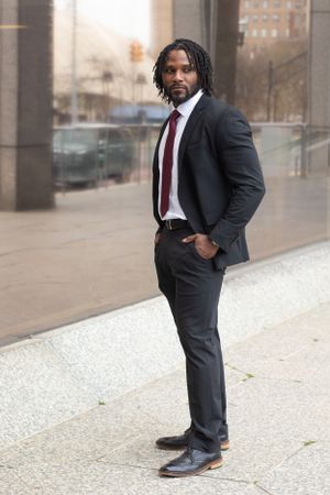 Professional male in dark suit and tie poses confidently against urban backdrop.