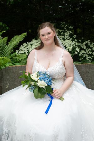 A bride in a white lace wedding gown holds a blue and white bouquet while seated outdoors with ferns and flowers behind.
