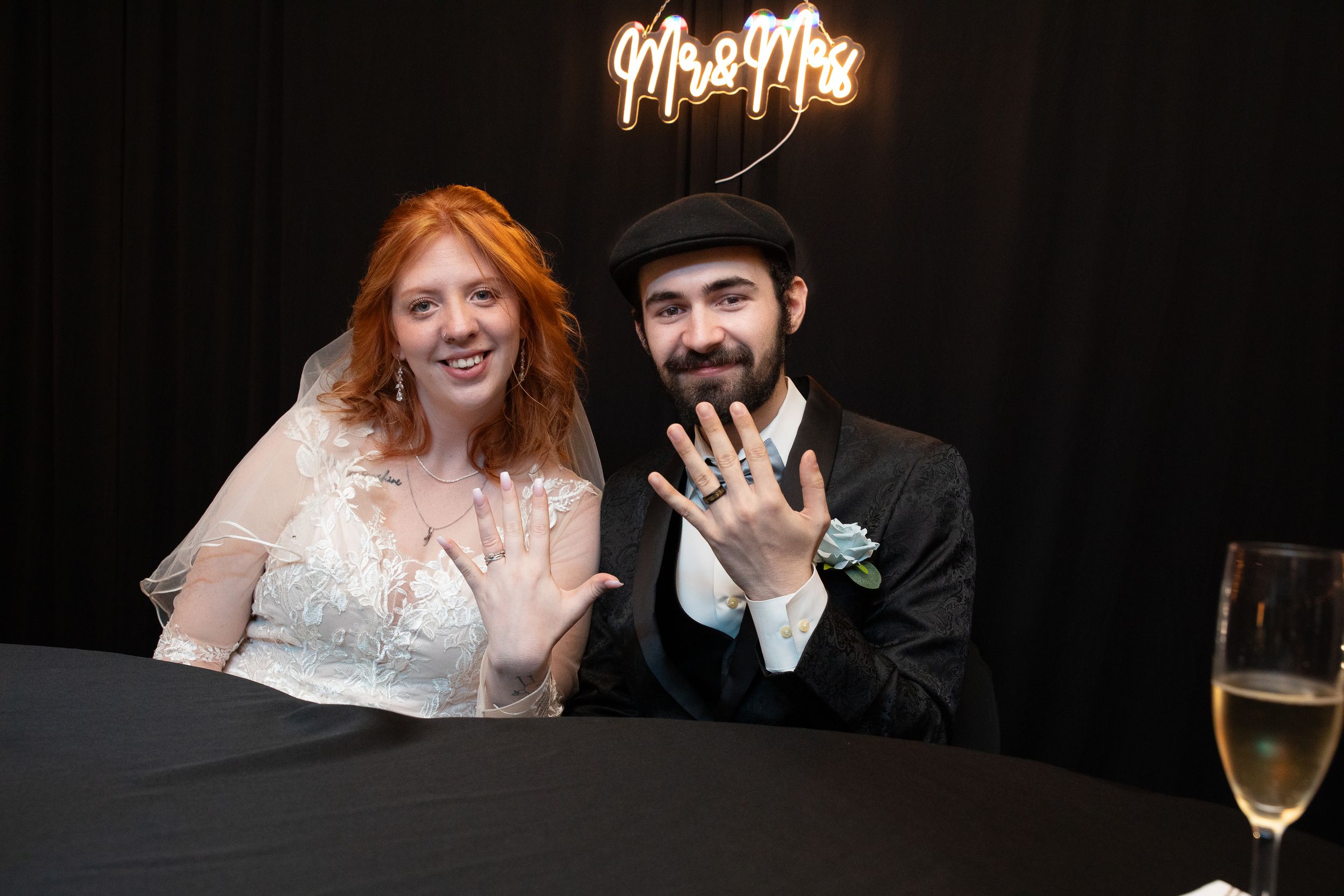Newlywed couple showing wedding rings at reception table under Mr & Mrs neon sign, bride in white veil, groom in black hat.
