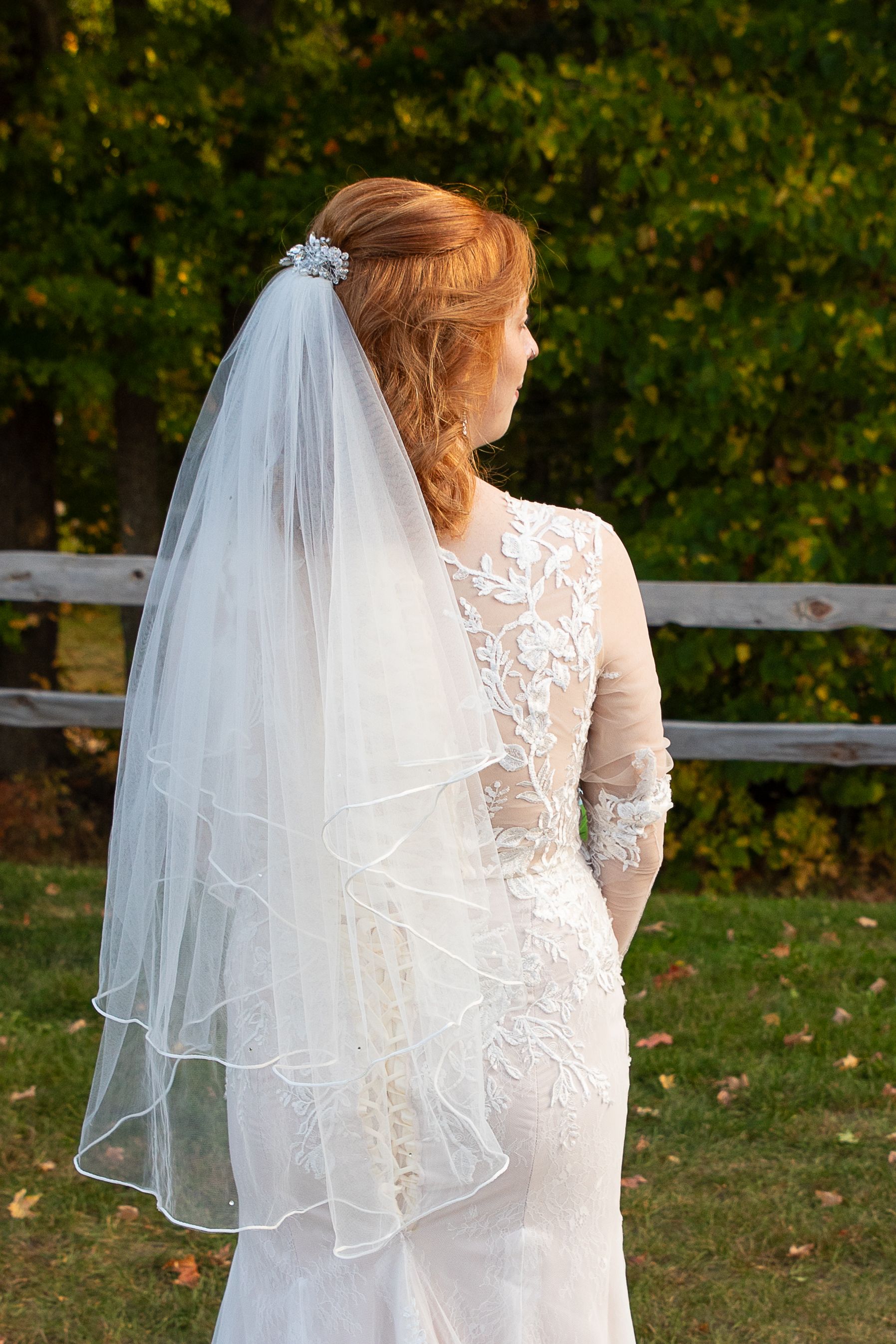 Bride in lace wedding gown and two-tier veil with beaded edge, viewed from behind outdoors.