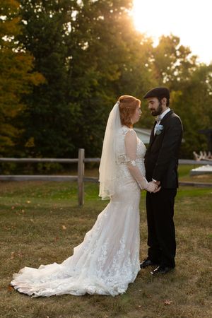 A bride and groom hold hands at golden hour, posing outdoors with lush green trees in the background.