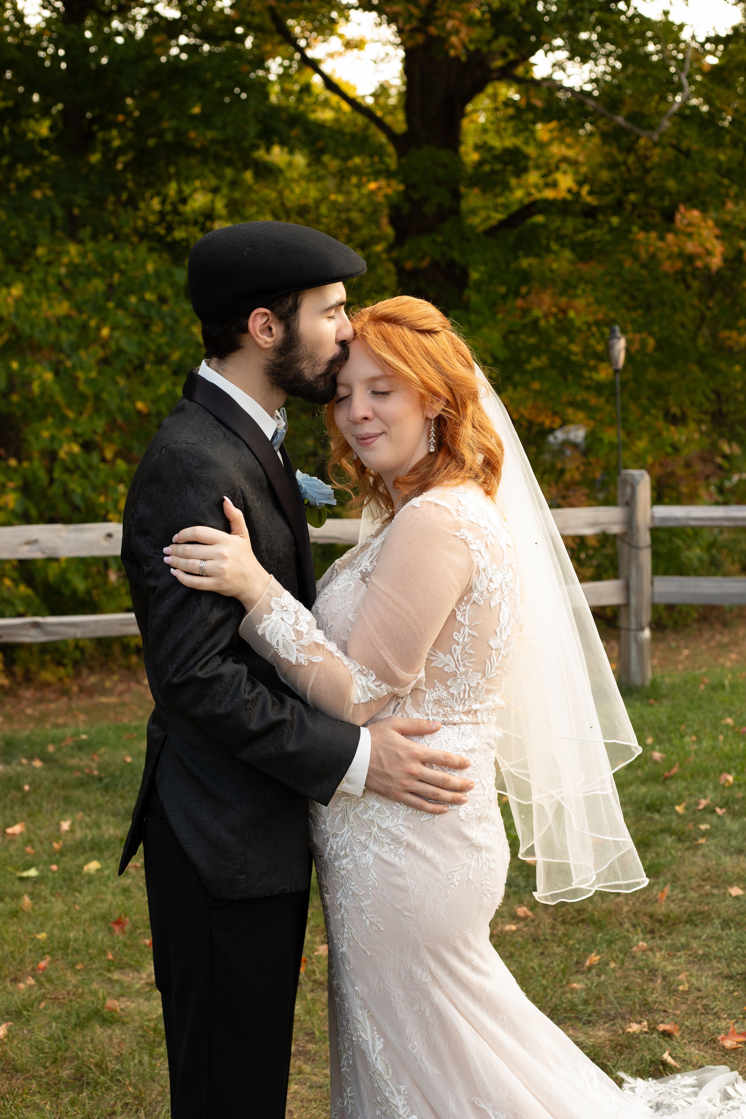 Bride in lace gown and veil embraces groom in black suit outdoors during autumn wedding portrait.