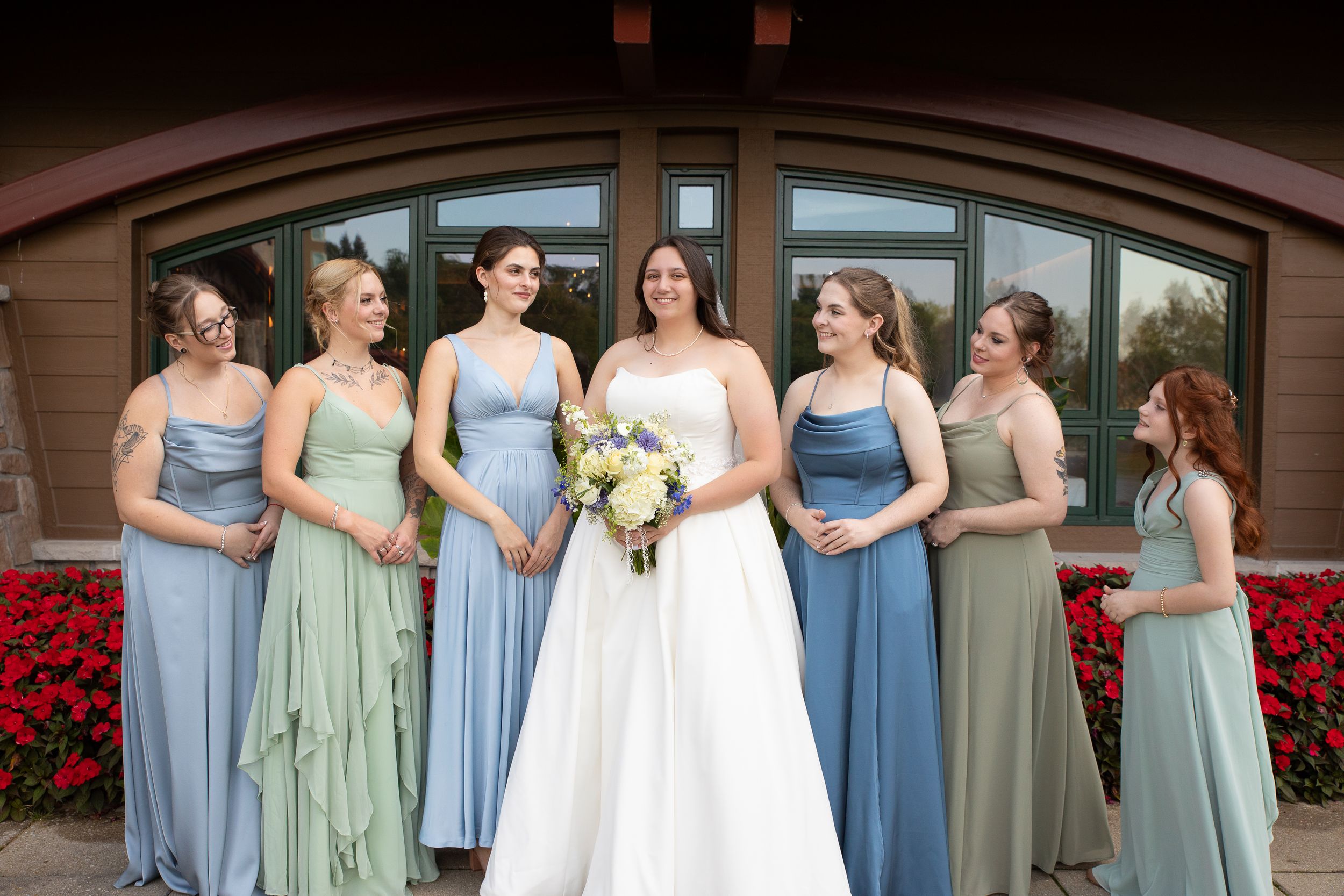Bride in white gown holds bouquet surrounded by six bridesmaids in sage green and blue dresses outside a rustic venue.