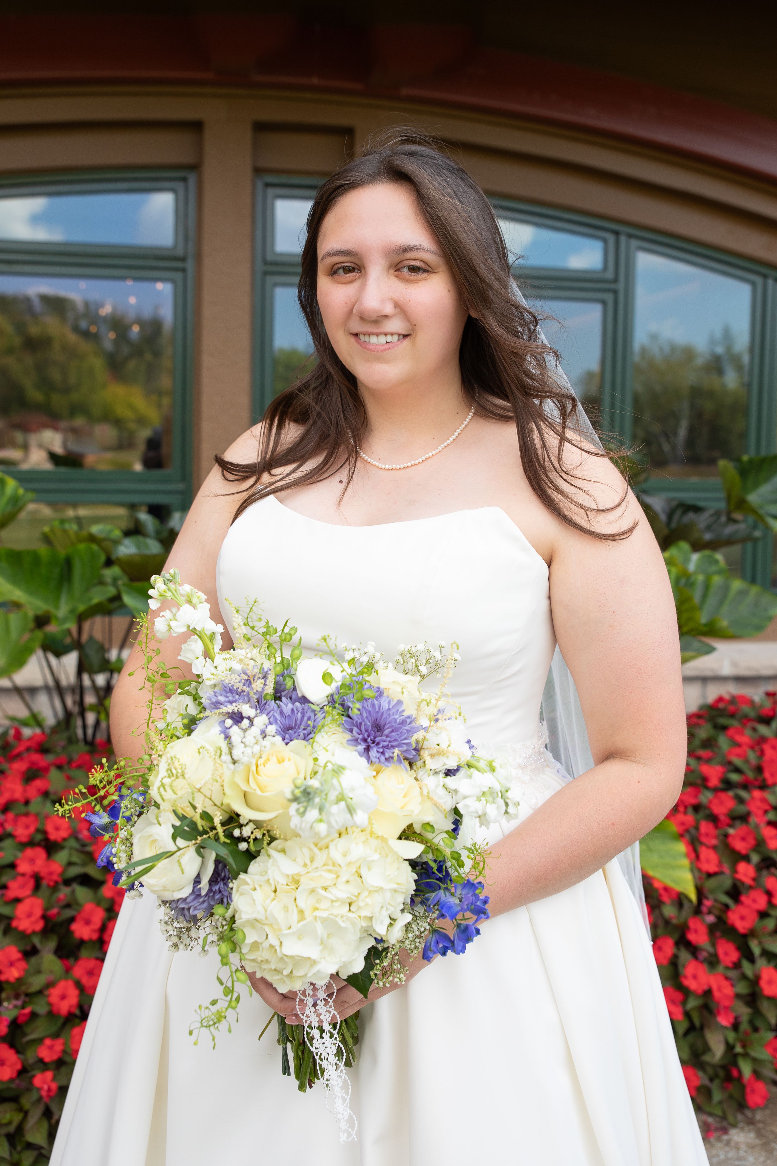Smiling bride in strapless white gown holding blue and white floral bouquet outdoors.