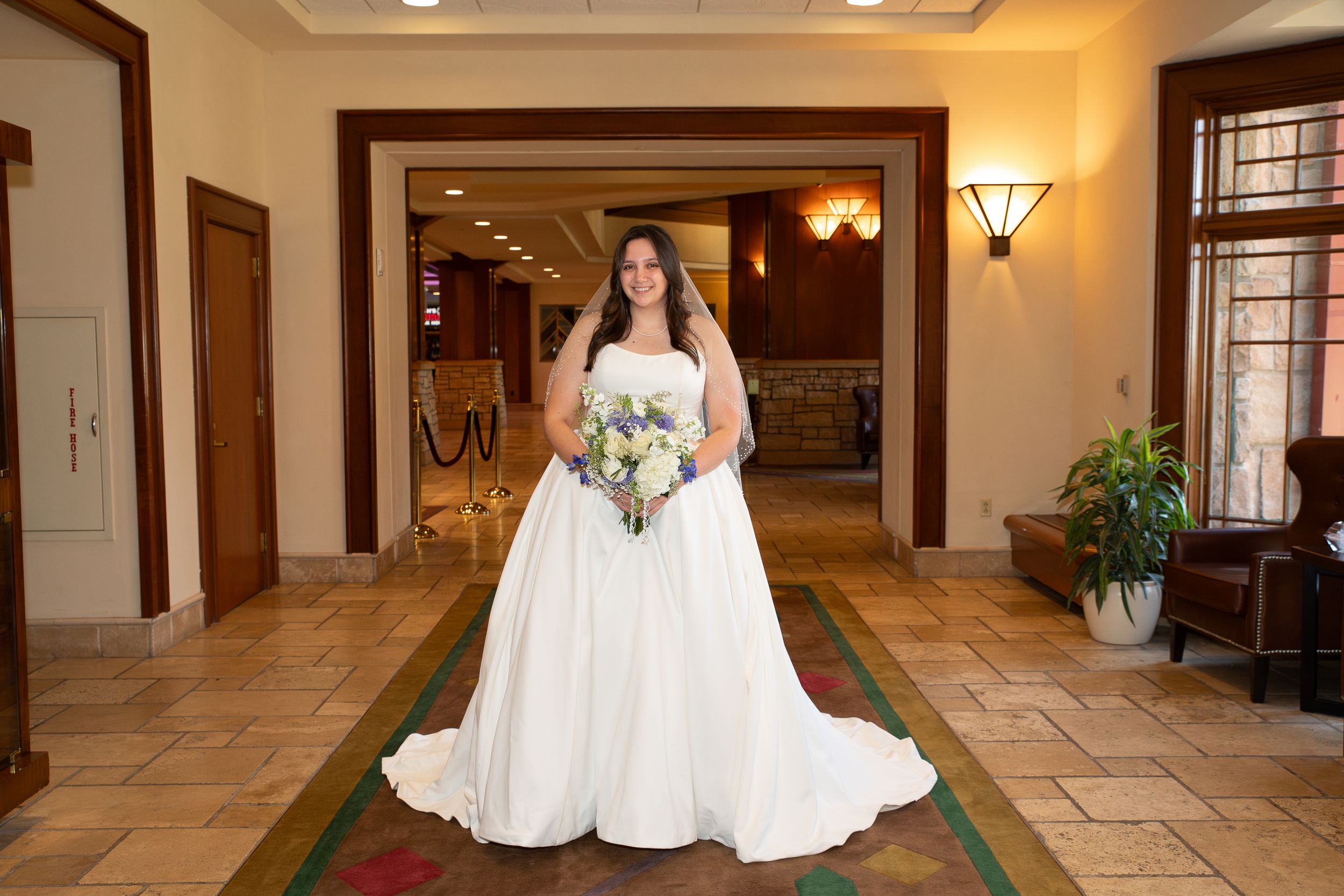 Bride in white ball gown holding bouquet stands in elegant venue lobby with warm lighting and decorative archways.