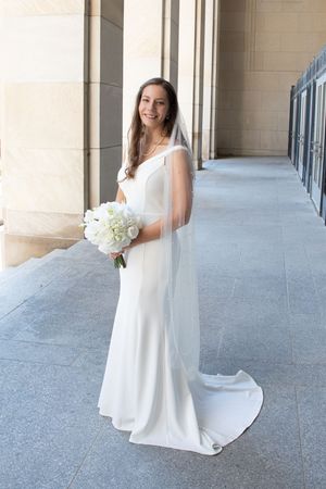 Bride in elegant white wedding gown holding white rose bouquet poses in columned hallway with natural lighting.