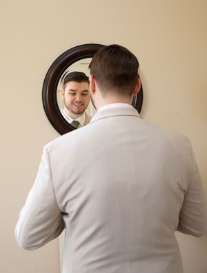 A person wearing a light-colored sweater looks in an oval-framed wall mirror while standing in a room with beige walls.
