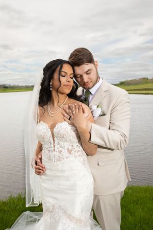 A romantic wedding couple embraces by a serene lake on their special day, with the bride in lace and groom in beige.