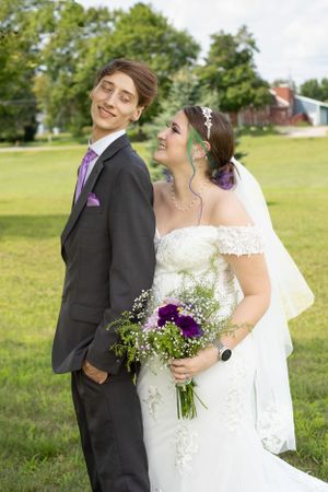 A wedding couple poses back-to-back outdoors with purple floral bouquet.