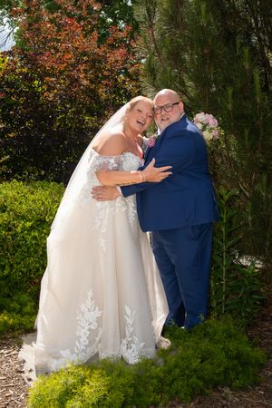 A joyful bride in a white lace gown embraces a bald groom in a blue suit outdoors among lush green garden shrubs.