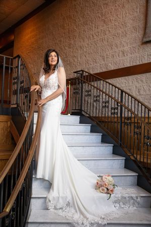 Bride in white lace gown with train stands on marble staircase holding railing, bouquet resting on steps behind her.