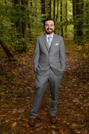 Groom in grey suit poses confidently on autumn forest path.