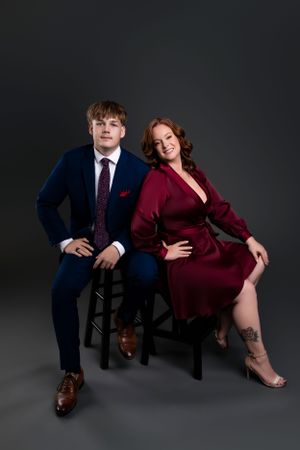 Professional studio portrait of two people seated on chairs against a dark background, wearing formal attire in navy and burgundy.