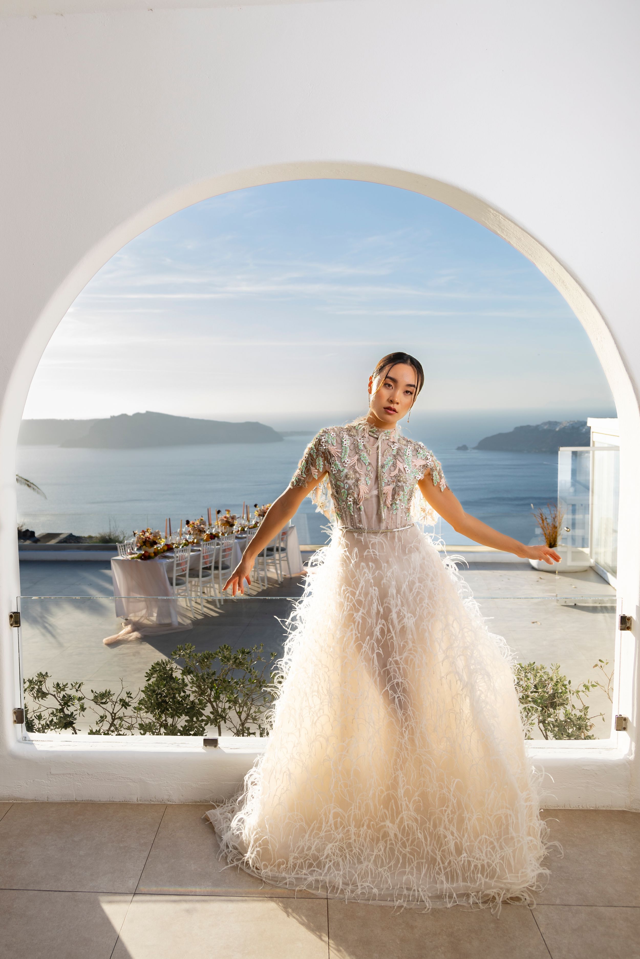 Luxurious white wedding gown with feathered skirt photographed against stunning Santorini ocean view through arched doorway.