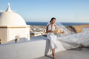 A person in a white dress poses against a white Mediterranean church dome overlooking coastal views.