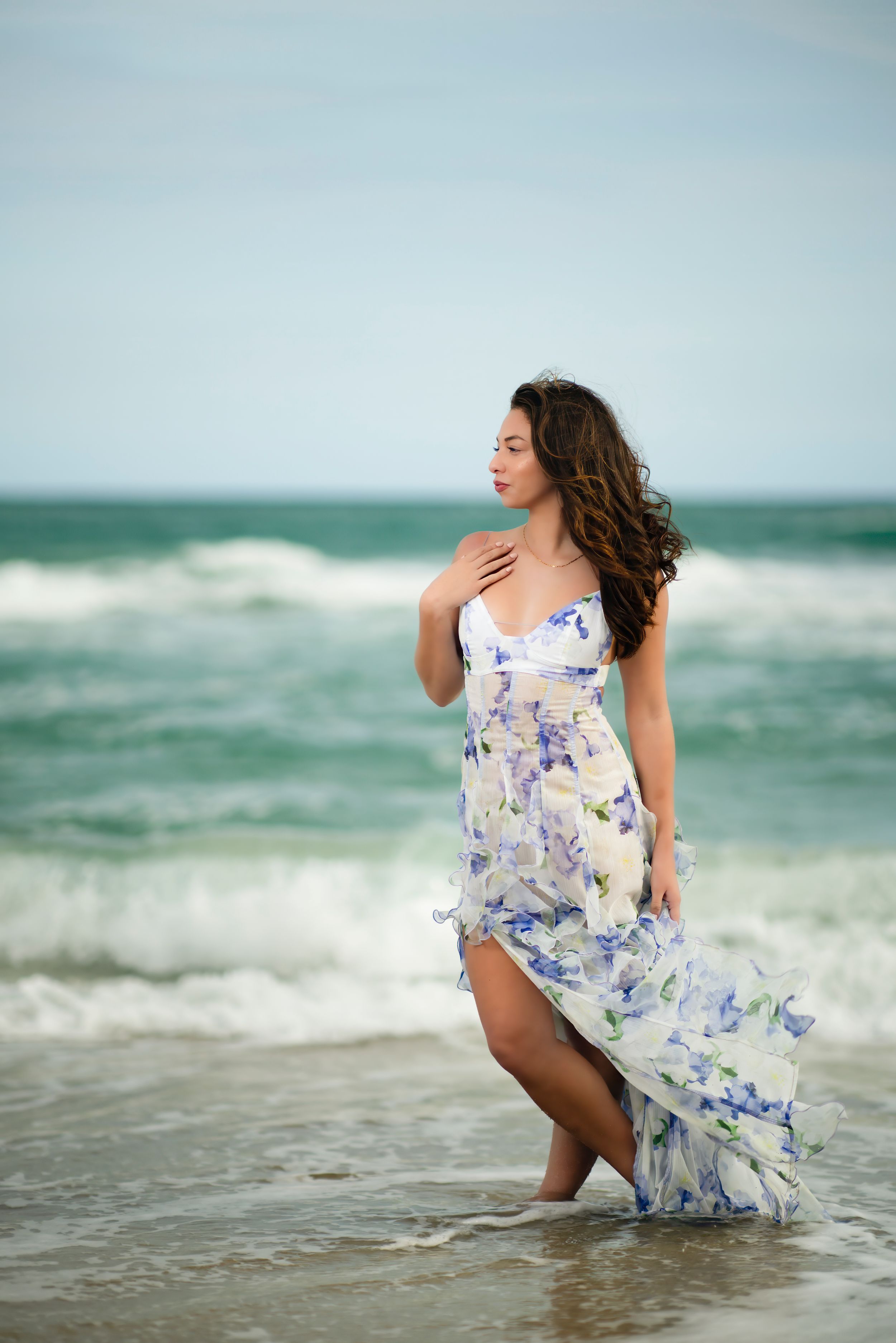 Woman in flowing white and blue floral dress poses gracefully at the beach with ocean waves in the background.
