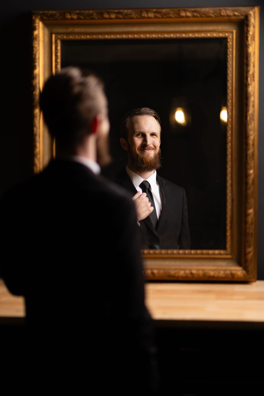 A person in a dark suit gazes at their reflection in a gilded framed mirror with warm lighting in the background.