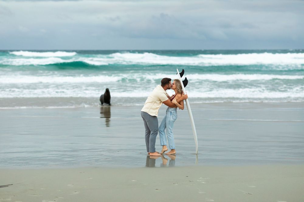 Two people embrace on a stormy beach while taking photos with a tripod as waves crash in the background.