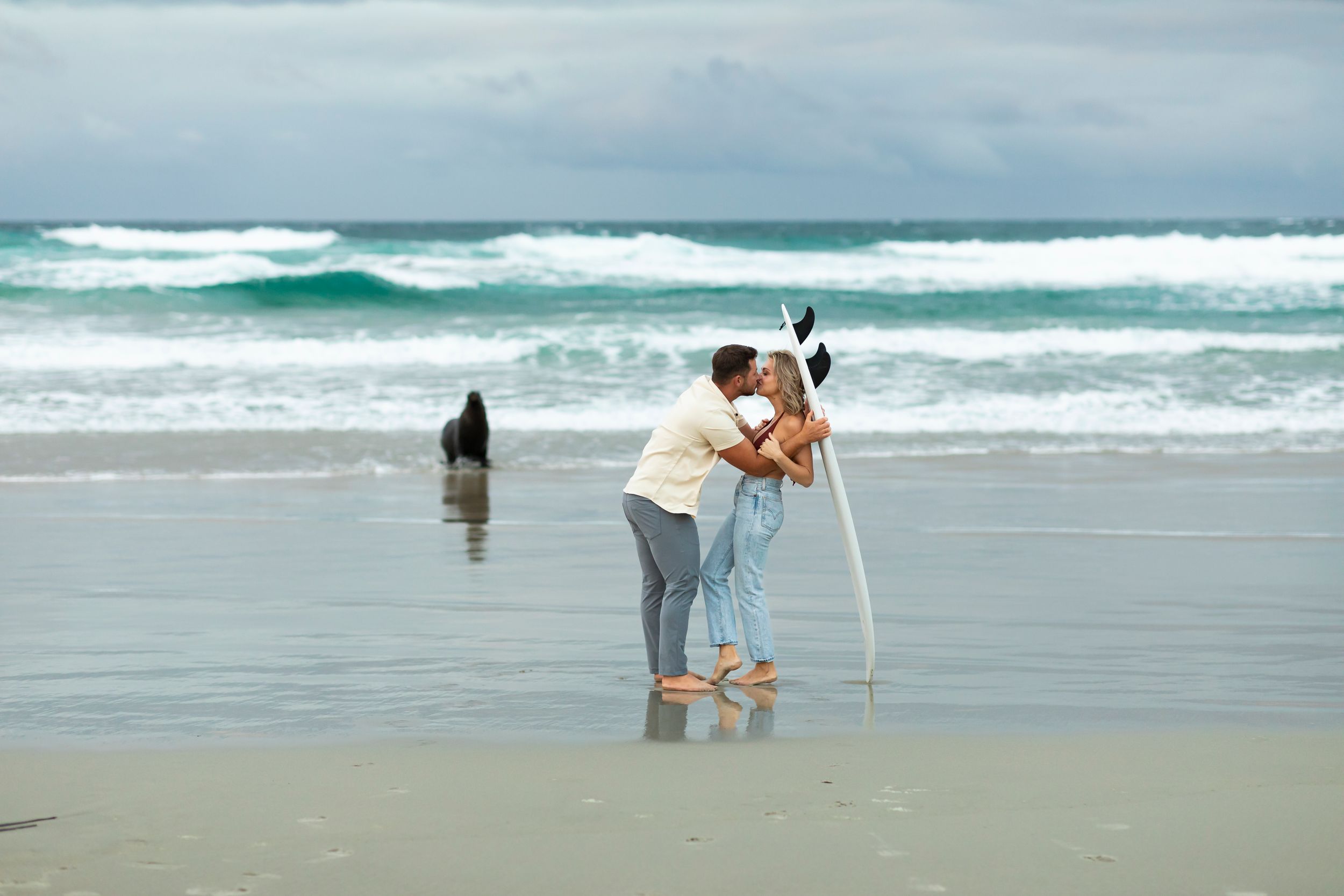 Two people embrace on a stormy beach while taking photos with a tripod as waves crash in the background.