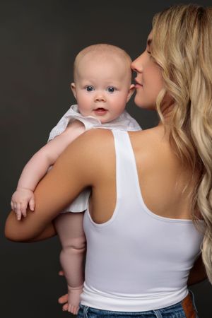 A mother in a white tank top holds and snuggles with a baby against a dark background.
