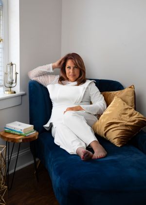 Person relaxing on a navy blue velvet couch with gold throw pillows next to a small side table with stacked books.