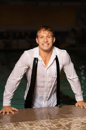 Someone wearing a wet white dress shirt and suspenders emerges from a swimming pool with a bright smile.