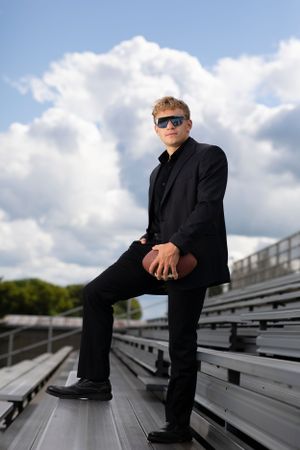Someone in a sharp black suit and sunglasses poses confidently on metal bleachers against a cloudy blue sky backdrop.