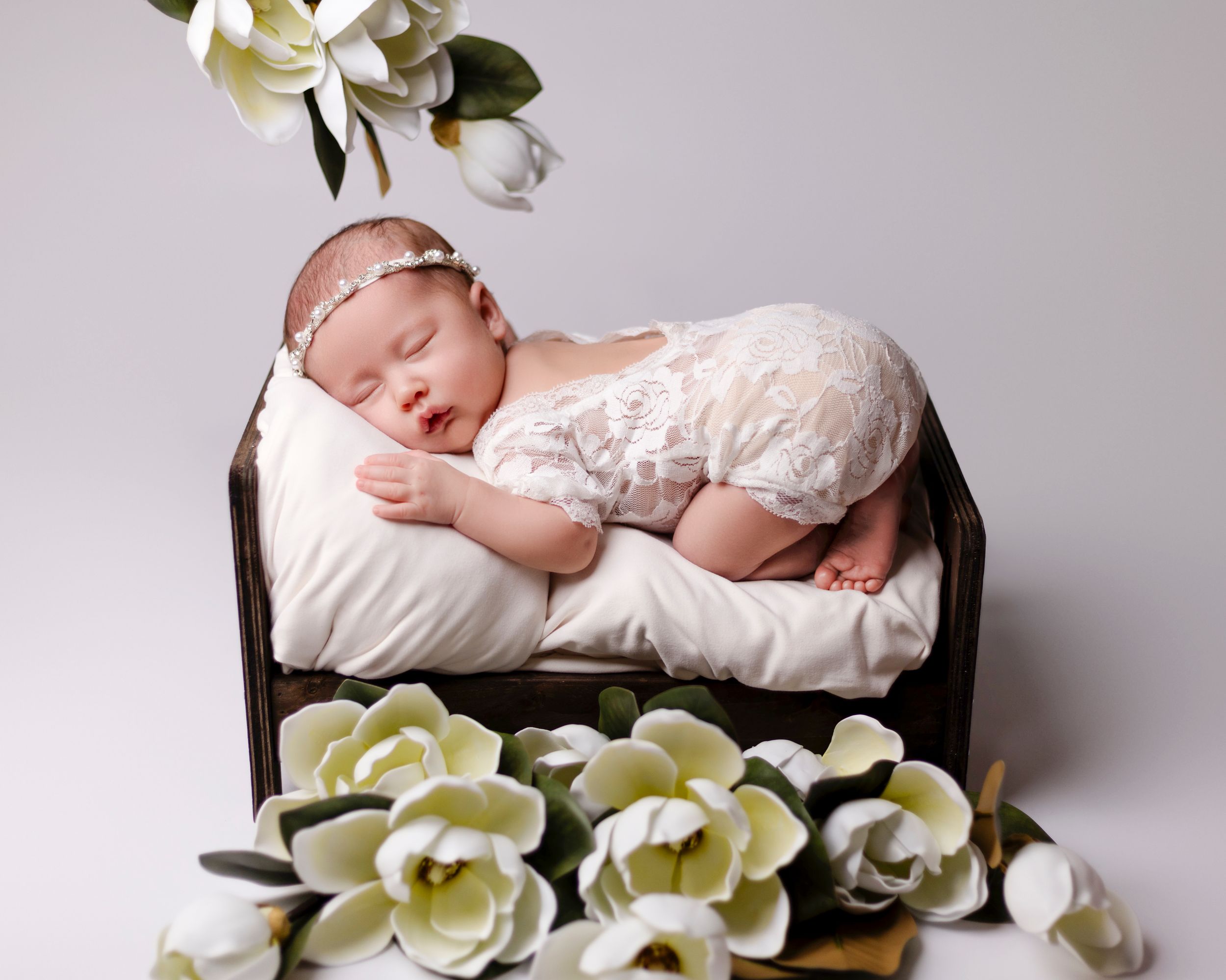 A newborn baby sleeping peacefully in a wooden basket adorned with soft white and yellow magnolia flowers.