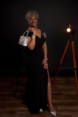 Elegant evening scene with silver-haired figure in black formal gown posing with purse near vintage tripod lamp on wooden floor.