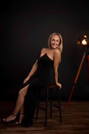 Elegant portrait of someone in a black evening gown seated on a bar stool against a dark backdrop with warm lighting.