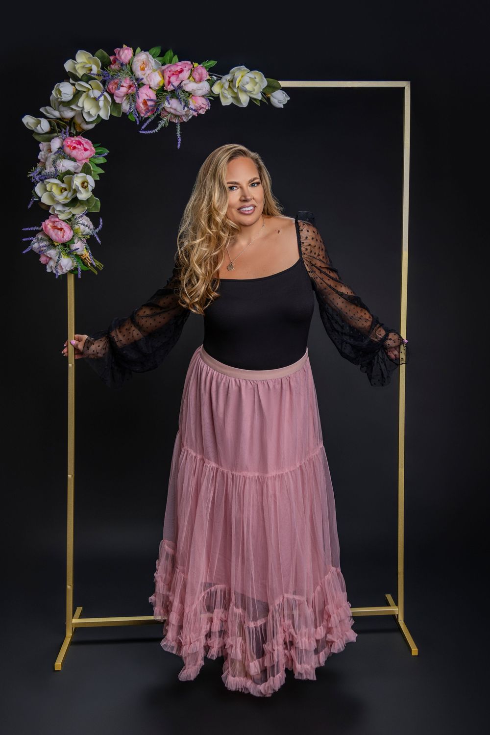A model poses in a gold frame with floral decor wearing a black top with sheer sleeves and flowing pink maxi skirt.