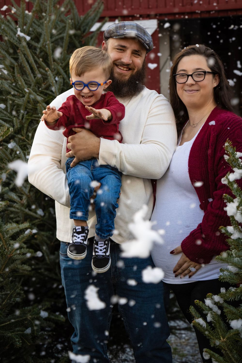 Winter family portrait with snow falling against evergreen trees and red barn background.
