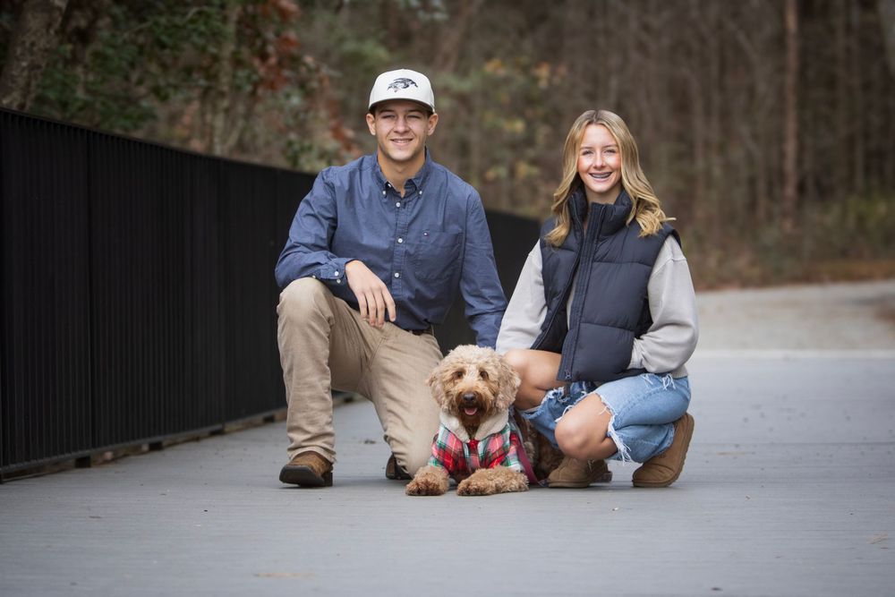 A couple sits with their small brown dog on a paved path next to a black fence in a wooded area.