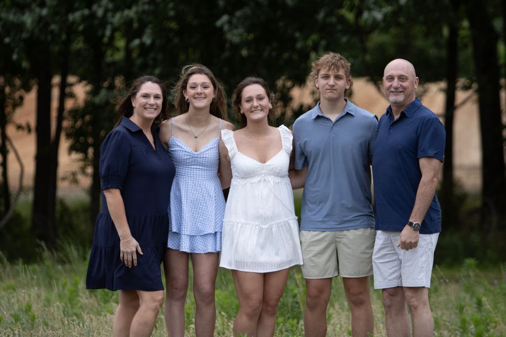 Family of five smiling outdoors in casual summer attire, posing together on a warm day with trees in background.
