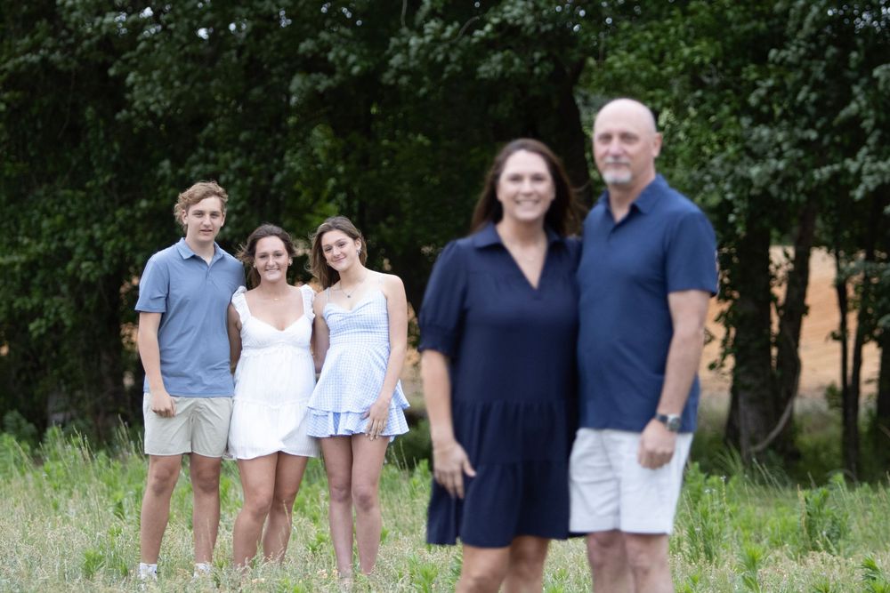 A family of five poses outdoors in a grassy field with trees, two adults in front and three teens behind.