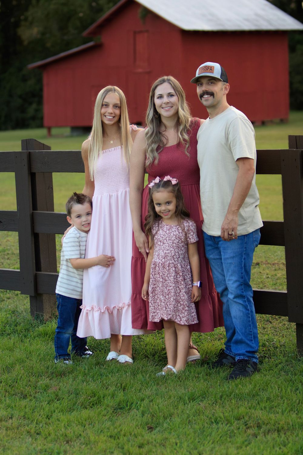 Family poses together in front of a red barn and wooden fence on a grassy lawn.