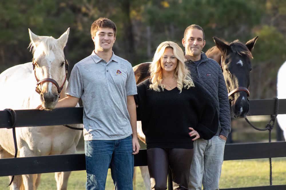 Family poses with their horses against a black fence at sunset in a rustic outdoor setting.