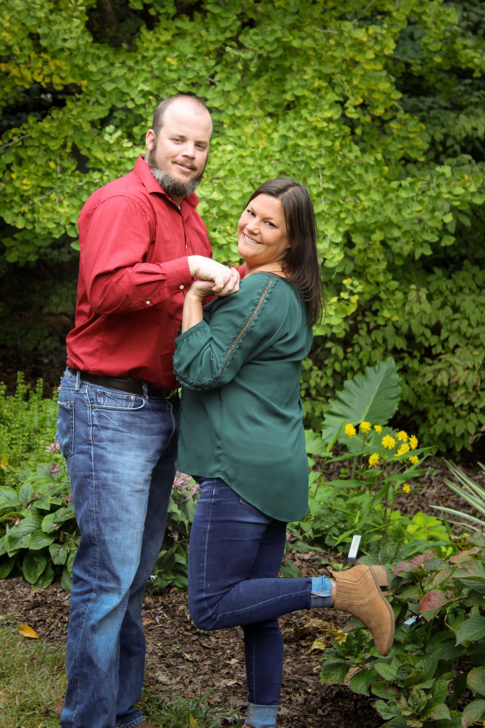 A couple in casual attire playfully poses together in front of green foliage in a garden setting.