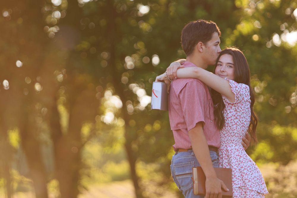 Young couple embracing in a sunlit park, woman in floral dress hugging man in pink shirt among green trees.