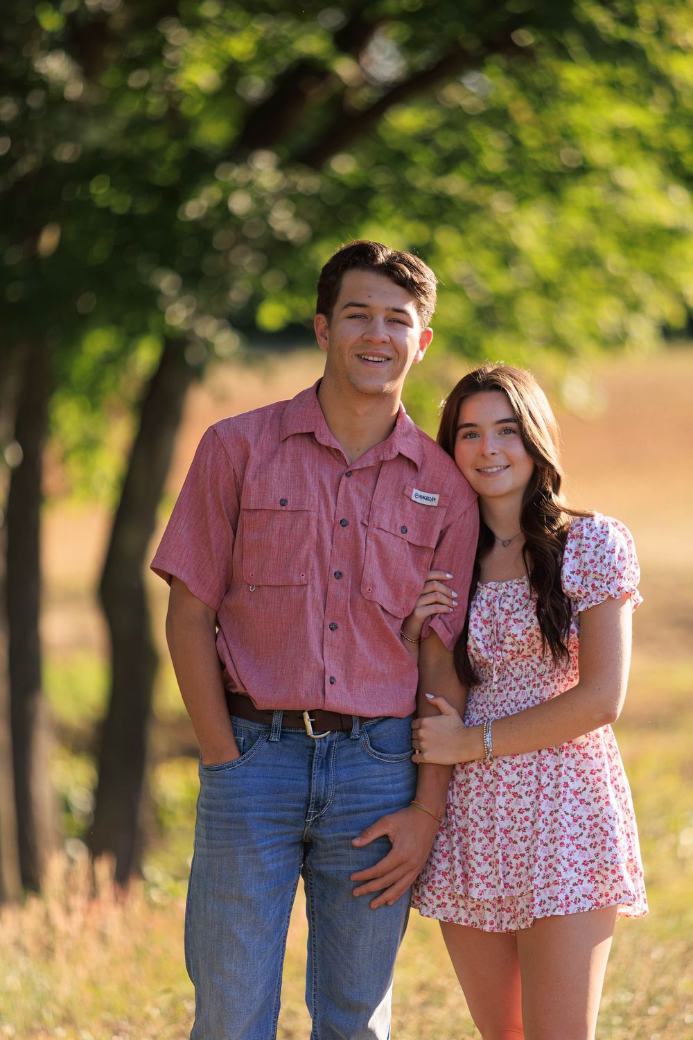 Young couple smiling outdoors in summer, man in red shirt and jeans, woman in floral dress, green trees background.