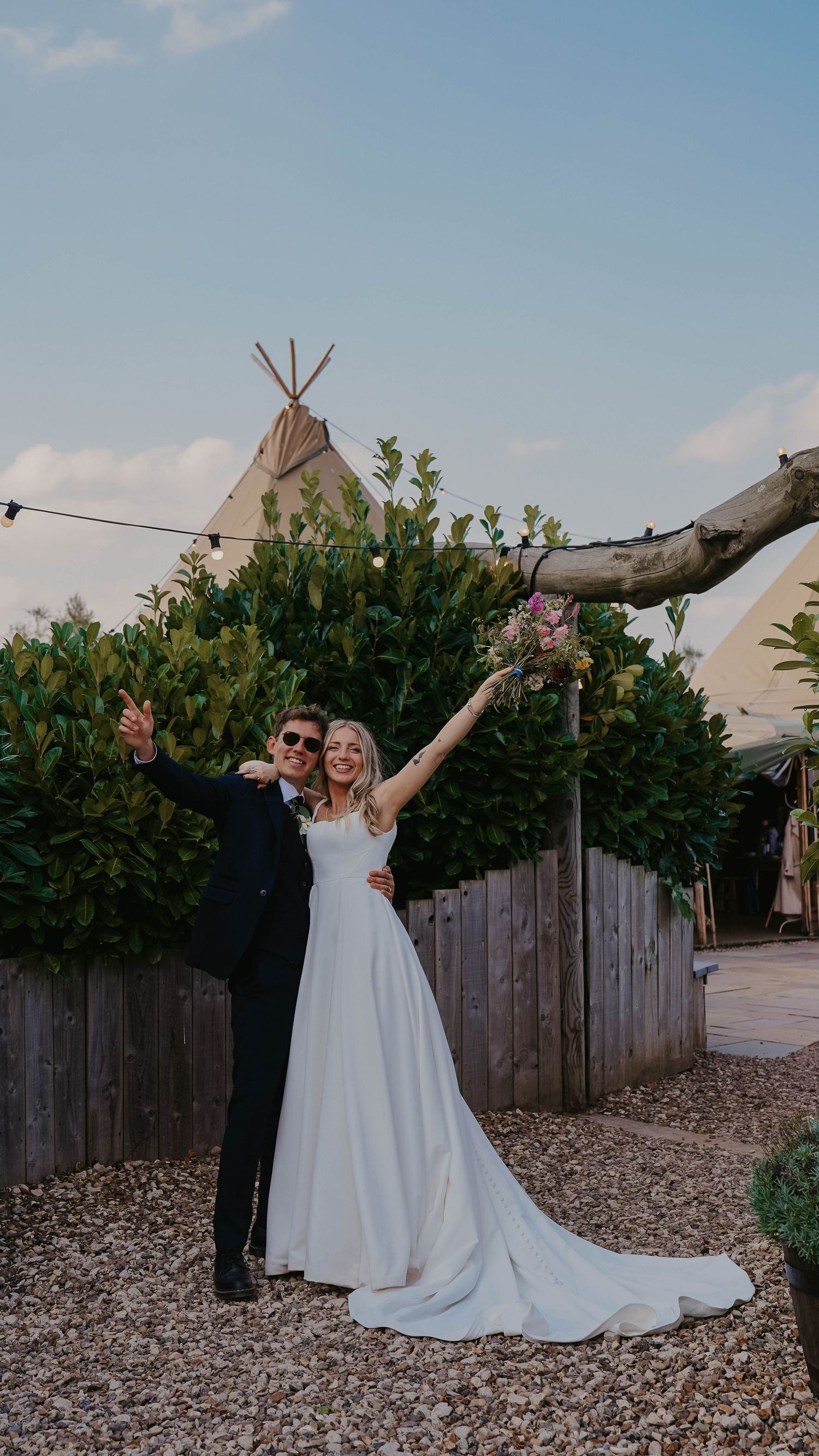Couple striking playful pose in front of teepee structure with flowing white dress train.