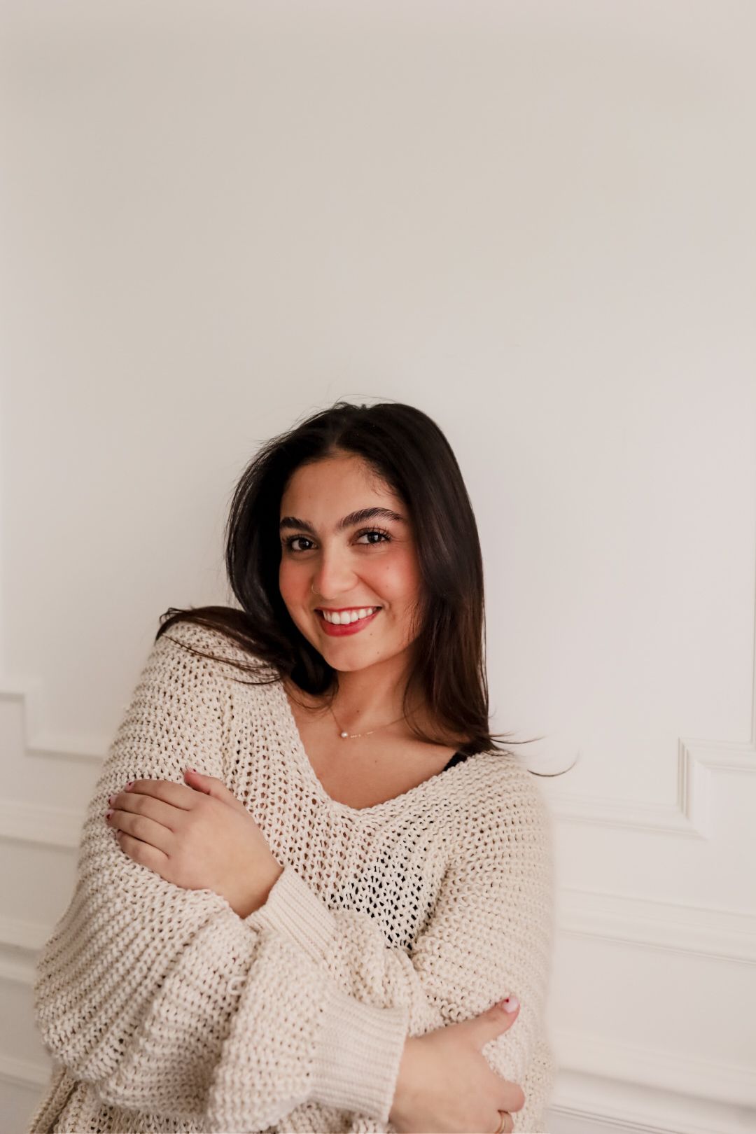 Brunette woman smiles warmly hugging herself in a cream knit sweater against a white wall.