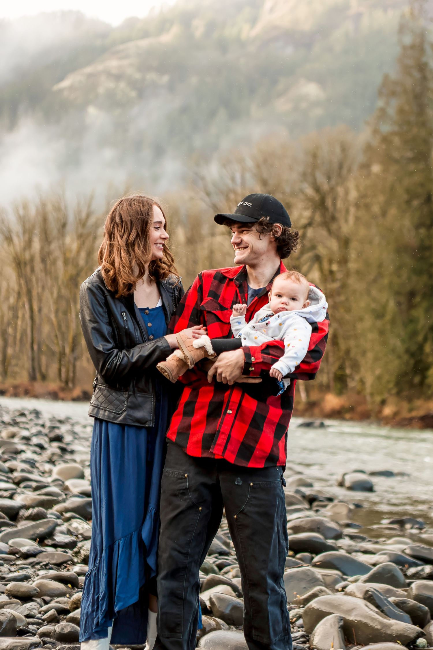 Family shares tender moments together on rocky riverbank with mountain backdrop and forest scenery.
