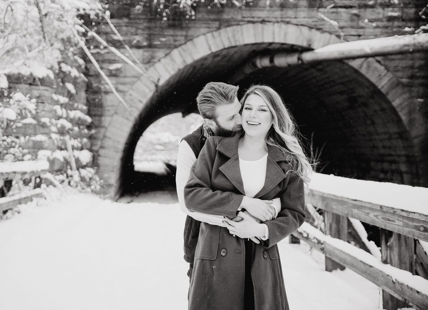 A black and white winter photograph showing a joyful moment between two people embracing on a snowy bridge near a stone arch.
