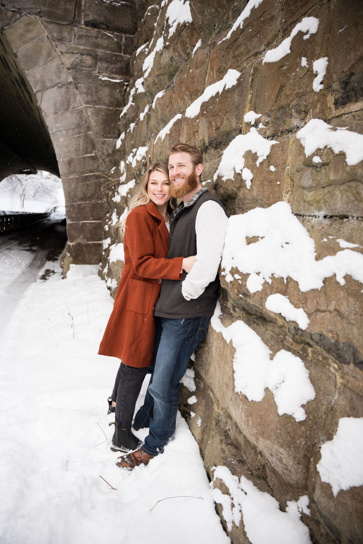 A couple enjoys a winter moment together beside a snow-covered stone wall under an archway.